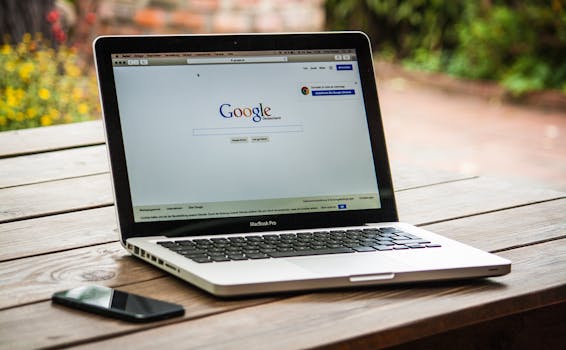 A MacBook Pro displaying Google Search on a wooden table outdoors, next to a smartphone.